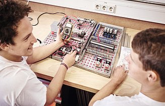 Foto zwei Arbeiter vor Schaltungsplatte, einer mit Messgerät in der Hand