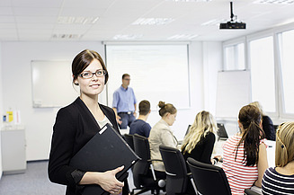 Frau steht mit Tablet in der Hand im Vordergrund, im Hintergrund sitzen Kolleg:innen in einem Konferenzraum.                  