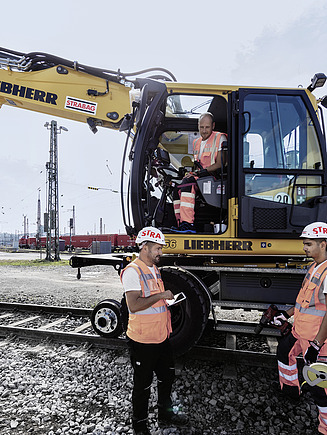 Foto drei Bauarbeiter besprechen sich vor einem Bagger, ein Vierter sitzt am Gerät