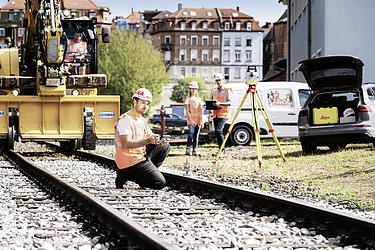 Foto Bauarbeiter kniet auf Gleisen, 2 Mitarbeitende im Hintergrund