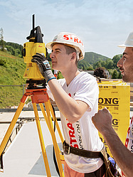 2 STRABAG Lehrlinge schauen auf ein Messgerät, das auf einem Stativ montiert ist.